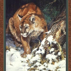 May include: A brown and tan cougar with green eyes stands in a snowy forest. The cougar is looking to the left of the image. The cougar is standing near a tree trunk covered in snow.