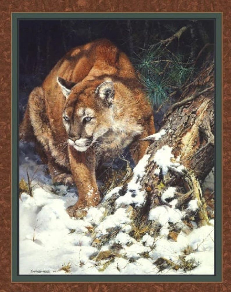 May include: A brown and tan cougar with green eyes stands in a snowy forest. The cougar is looking to the left of the image. The cougar is standing near a tree trunk covered in snow.
