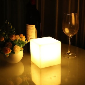 May include: A white cube-shaped LED lamp with a warm white glow, sitting on a table with a bottle of wine and a wine glass.