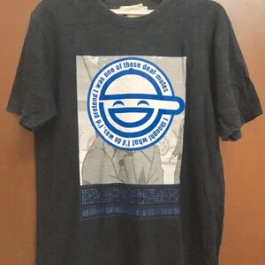 Könnte beinhalten: Dunkelgraues T-Shirt mit einem Motiv aus der Anime-Serie Ghost in the Shell. Das Design zeigt ein lächelndes Gesicht in einem blauen Kreis, mit englischem Text. Das T-Shirt hängt an einem weißen Kleiderbügel.