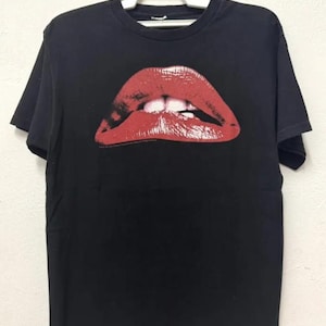 Puede incluir: Camiseta negra con un gran estampado de labios rojos. Los labios est&aacute;n abiertos y muestran los dientes.