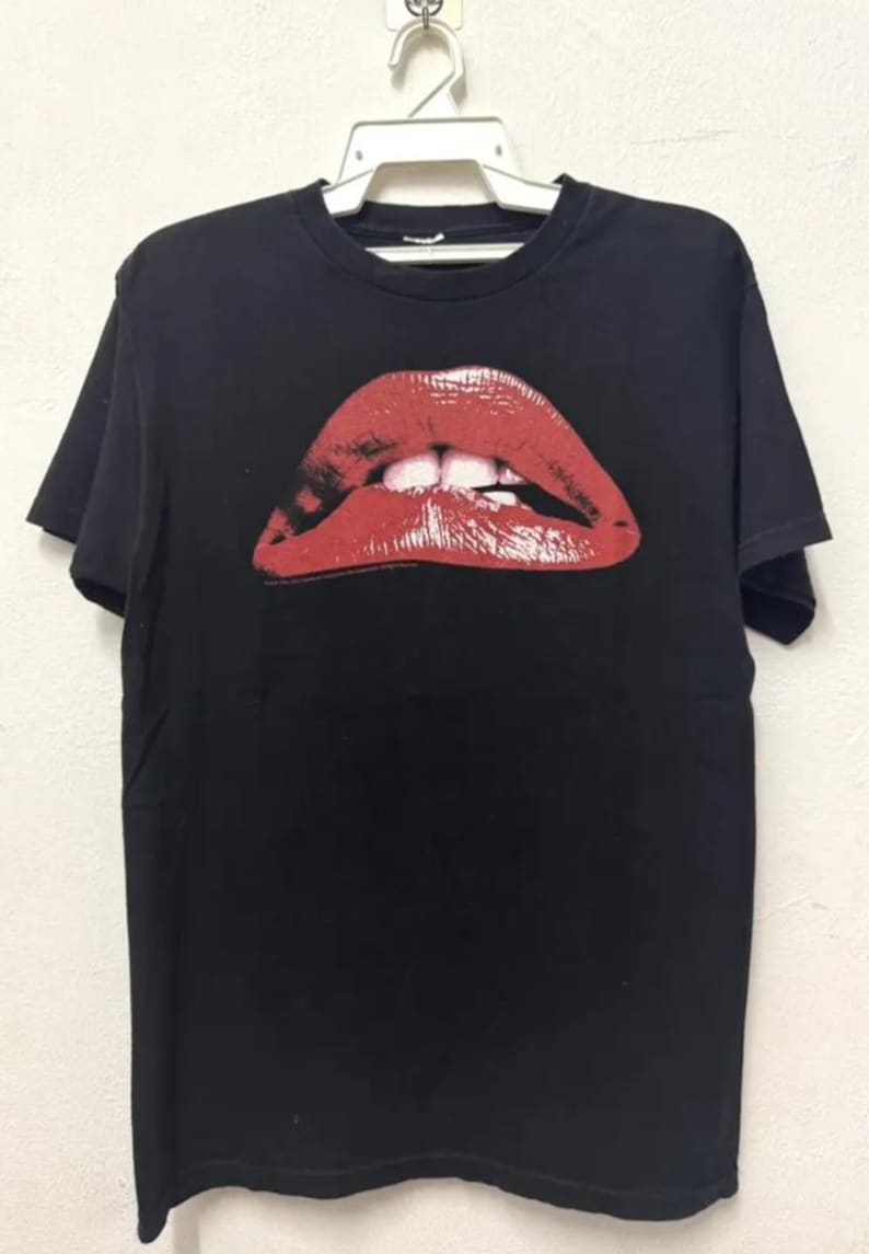 Puede incluir: Camiseta negra con un gran estampado de labios rojos. Los labios est&aacute;n abiertos y muestran los dientes.