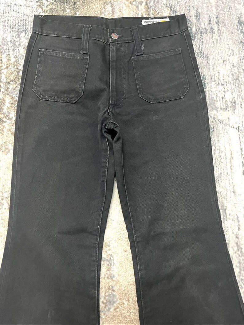 Może przedstawiać: Czarne dżinsy Wrangler z denimu z wysokim stanem i rozszerzaną nogawką. Dżinsy mają dwie przednie i dwie tylne kieszenie. Logo Wrangler jest widoczne na tylnej kieszeni. Dżinsy są wykonane z wytrzymałego denimu.