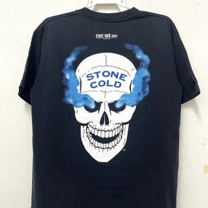 May include: Black t-shirt featuring a white skull graphic. The skull has blue smoke emanating from its eye sockets. The text "STONE COLD" is on the skull's forehead. The text "STONE COLD.com" is printed on the shirt.