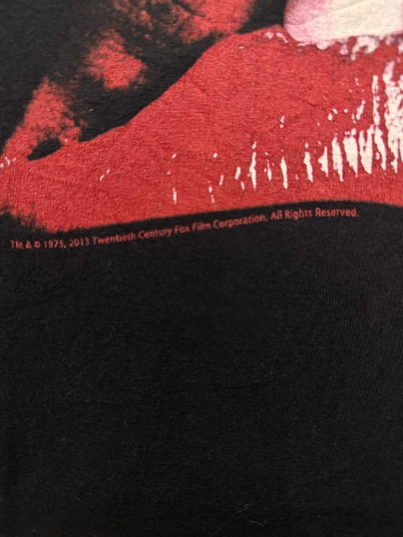 Puede incluir: Una camiseta negra con una imagen roja y blanca de un par de labios. El texto "TM & &copy; 1975, 2013 Twentieth Century Fox Film Corporation, All Rights Reserved." est&aacute; impreso en la parte inferior de la camiseta.