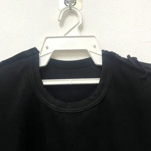 Può includere: Una maglietta nera appesa a un appendiabiti di plastica bianca. La maglietta ha un collo rotondo e maniche corte.