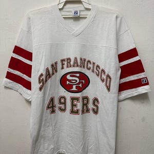 Könnte beinhalten: Weißes V-Ausschnitt-T-Shirt mit roten Streifen auf den Ärmeln. Das Shirt zeigt die Worte "SAN FRANCISCO" und "49ERS" in Braun und Rot, mit dem 49ers-Logo in der Mitte.