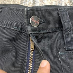 Może przedstawiać: Zbliżenie na ciemnoszare dżinsy z guzikiem z czerwonym logo "Wrangler". Złoty zamek błyskawiczny jest otwarty, odsłaniając granatowy rozporek. Spodnie są lekko pomarszczone.
