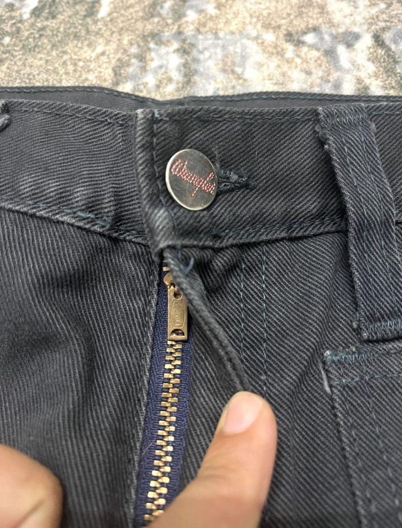 Może przedstawiać: Zbliżenie na ciemnoszare dżinsy z guzikiem z czerwonym logo "Wrangler". Złoty zamek błyskawiczny jest otwarty, odsłaniając granatowy rozporek. Spodnie są lekko pomarszczone.