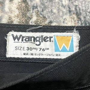 Może przedstawiać: Zbliżenie czarnego elementu garderoby z dżinsu z białą etykietą. Na etykiecie widnieje napis "Wrangler JEANS & SPORTSWEAR" oraz "ROZMIAR 30 IN / 76 CM". Logo Wrangler to ż&oacute;łto-niebieskie stylizowane "W".