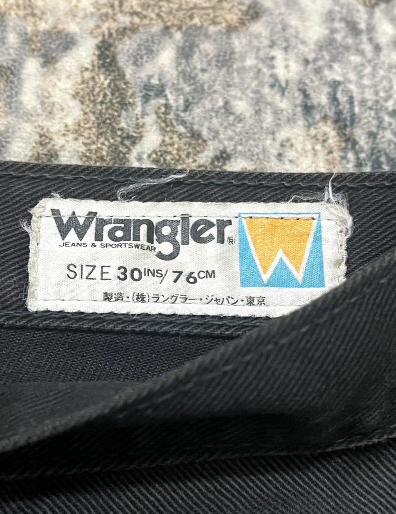 Może przedstawiać: Zbliżenie czarnego elementu garderoby z dżinsu z białą etykietą. Na etykiecie widnieje napis "Wrangler JEANS & SPORTSWEAR" oraz "ROZMIAR 30 IN / 76 CM". Logo Wrangler to ż&oacute;łto-niebieskie stylizowane "W".