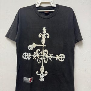 以下が含まれることがあります： 白い装飾的な十字のデザインが特徴の黒いTシャツ。左下に小さな「ローリング・ストーンズ」のパッチがあります。Tシャツは白いハンガーに吊るされています。