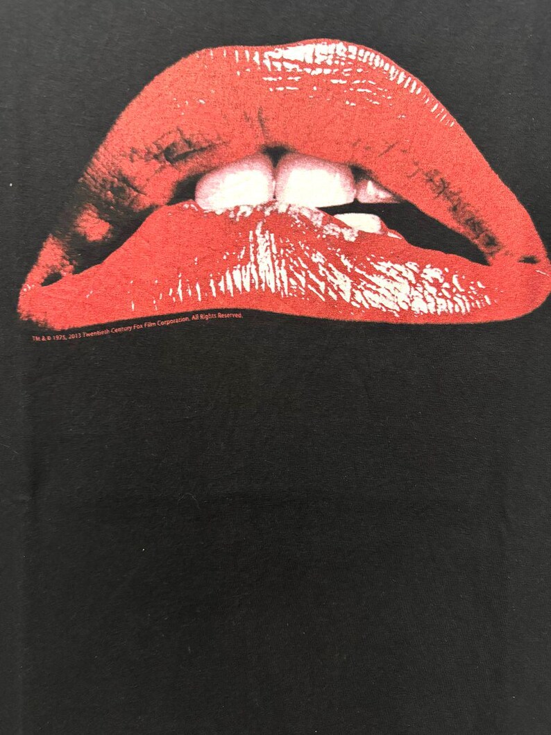Puede incluir: Una camiseta negra con una imagen roja, blanca y negra de un par de labios. Los labios est&aacute;n abiertos y los dientes son visibles. El texto "TM & &copy; 1982, 2012 Twentieth Century Fox Film Corporation. All Rights Reserved." est&aacute; impreso en la parte inferior de la imagen.