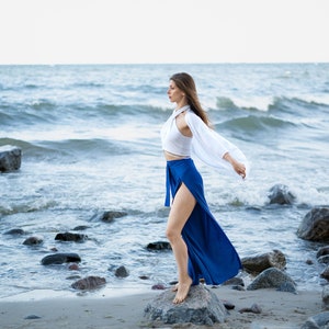 tall model in cobalt blue high slit pants, white crop top and white kimono, standing on a rock by the sea, the clothes are flowing in the wind