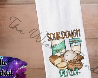 May include: White kitchen towel with a colorful illustration of sourdough bread, flour, and a jar of starter. The text "SOUR DOUGH DEALER" is printed on the towel in brown and teal lettering. The towel is displayed on a wooden surface.