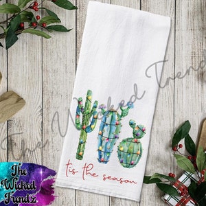 May include: A white kitchen towel with a Christmas cactus design. The cactus is decorated with green and red Christmas lights. The text "Tis the season" is printed in red on the towel.