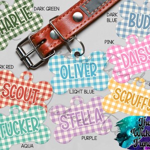 May include: A collection of dog tags in various colors and patterns, including dark green, dark blue, pink, orange, aqua, and purple. Each tag features a different name, such as "Charlie," "Buddy," and "Daisy," in a playful font. A brown leather dog collar is also visible.