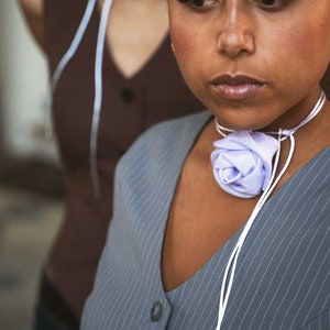 May include: A woman wearing a gray pinstripe top with a white ribbon tied around her neck. The ribbon has a light purple fabric flower tied in the center.