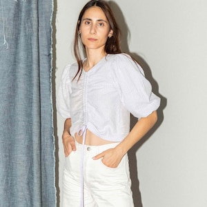 Può includere: Una donna indossa una blusa bianca corta e attillata con un laccio in lavanda e pantaloni bianchi larghi.
