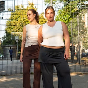 May include: Two people wearing matching outfits. One person is wearing a cream-colored tank top and brown pants. The other person is wearing a white tank top and dark gray pants. They are standing in front of a chain-link fence.