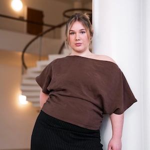 Puede incluir: Una mujer con un top marrón con hombros descubiertos y pantalones negros de raya diplomática. El top tiene mangas cortas y un diseño drapeado. Los pantalones son de cintura alta. El fondo presenta una columna blanca y una escalera de caracol.