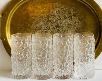 May include: Four clear glass tumblers with a textured, frosted surface. The glasses are arranged in a row on a white surface.