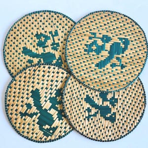 May include: Four round woven coasters. The coasters are made of natural-colored straw with a dark teal geometric design in the center. The edges are trimmed with a dark teal border.