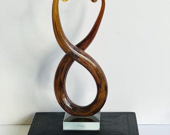 May include: A decorative glass sculpture with a swirling infinity symbol design. The sculpture is primarily brown with gold accents at the top. It is mounted on a clear, rectangular base. The sculpture is displayed on top of a dark book.
