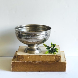 May include: A silver-toned metal bowl with a pedestal base, resting on two stacked, aged books. The bowl has a decorative rim and the inscription "family is belonging to and becoming". A small green plant adds a touch of color.