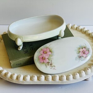 May include: An oval, off-white ceramic soap dish with three small feet, and a matching lid decorated with pink floral designs. The dish and lid rest on a cream-colored, beaded tray, with a green book underneath.
