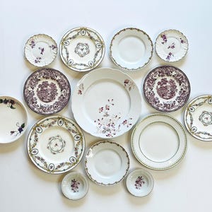 May include: A collection of antique decorative plates in various sizes and designs. The plates feature floral patterns, intricate borders, and a mix of colors including white, purple, and green. The arrangement is on a white background.