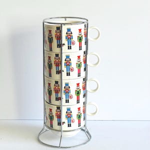 May include: A set of four white ceramic mugs stacked vertically in a chrome metal wire rack. Each mug features a repeating pattern of colorful nutcracker figures in red, blue, green, and black uniforms, along with small red snowflake-like accents.