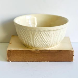 May include: A cream-colored ceramic mixing bowl with a diamond pattern and vertical accents. The bowl sits on a light brown rectangular base. The bowl has a wide rim and a smooth interior.
