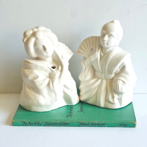 May include: Two white ceramic figurines of a woman and a man in traditional Japanese clothing. The woman is holding a fan and the man is holding a fan. The figurines are sitting on a green book titled "The Art of the Japanese Garden" by Tatsuo Ishimoto.