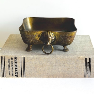 May include: A vintage brass bowl with a lion's head ring handle and floral detailing. The bowl is rectangular with curved sides and rests on four small feet. The bowl is placed on top of a book titled "The Complete Encyclopedia of Antiques."