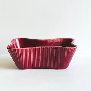 May include: A burgundy-colored ceramic bowl with a unique, wavy rim. The bowl has vertical, ribbed detailing on the exterior, adding texture to the design. The bowl is a decorative piece, suitable for serving or display.