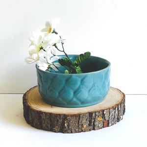 May include: A teal ceramic bowl with a textured pattern, holding white flowers and greenery, sits on a rustic wooden slice. The bowl's design adds a decorative touch, perfect for displaying floral arrangements.