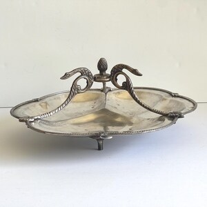 May include: A silver-toned metal serving dish with a three-sectioned tray design. The center features a vertical handle topped with a small pinecone finial, flanked by two curved, swan-shaped supports. The dish stands on three small feet.
