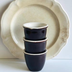 May include: A stack of three dark brown ceramic ramekins with white rims and interiors, set in front of a large, off-white, scalloped-edge plate. The ramekins are stacked, showcasing their rounded shape and contrasting colors.