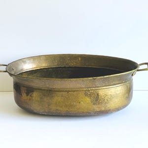 May include: An oval-shaped brass basin featuring a weathered, golden-brown patina and a smooth, rounded rim. The vessel includes two small, curved metal handles attached to opposite sides, providing a functional grip for this metallic container.