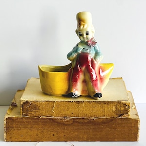 May include: Vintage ceramic planter featuring a boy figure in red overalls and a blue shirt, seated on a yellow boat-shaped planter. The planter is placed on top of a stack of old books with aged, yellowed pages and a wooden base.