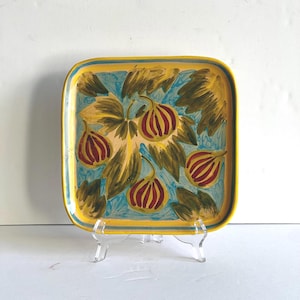 May include: Square ceramic serving tray with a hand-painted fig design. The tray features a yellow border, a light blue background, and a fig motif in red, yellow, and green. The tray is displayed on a clear acrylic stand.