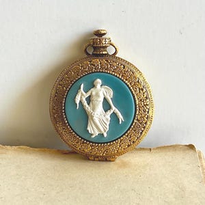 May include: A vintage gold-toned compact with a teal center featuring a white bas-relief of a classical figure holding a garland. The compact has a floral border and a decorative top loop.