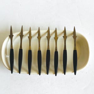 May include: A set of eight vintage appetizer forks with black handles and brass-colored spear-shaped tines. The forks are arranged on a white, oval-shaped serving dish, perfect for a retro-themed dinner party.