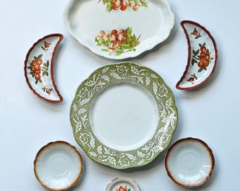 May include: A collection of vintage ceramic plates and dishes. The set includes a large oval platter with a fruit design, a green and white patterned plate, and several smaller plates and dishes with various designs and colors.