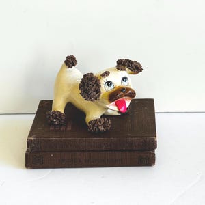 May include: A vintage ceramic dog figurine with a cream-colored body, brown accents, and a pink tongue. The dog is posed on top of two stacked, dark brown books. The figurine has a playful expression and is a decorative item.