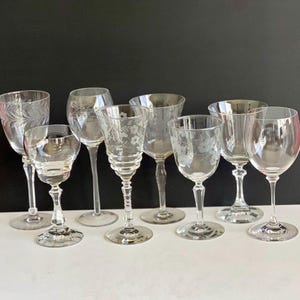 May include: A collection of clear, etched glass wine glasses. The glasses vary in shape and size, with floral and leaf patterns. Some have tall stems and rounded bowls, while others have shorter stems and wider bowls. The glasses are arranged on a white surface against a black background.