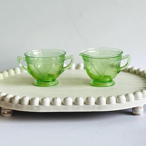 May include: Two green glass serving pieces, a sugar bowl with handles and a creamer, sit on a decorative tray. The pieces have a detailed pattern and are made of a light green glass. The tray is white with a beaded edge.
