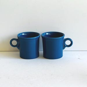 May include: Two cobalt blue ceramic mugs with rounded handles. The mugs have a cylindrical shape with a slightly flared rim. The mugs are displayed on a white surface. The mugs are a solid color and have a glossy finish.
