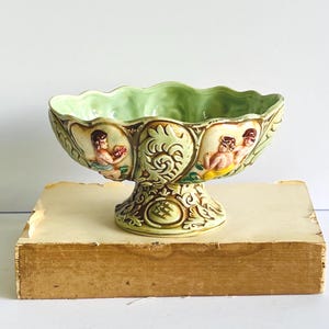 May include: A ceramic pedestal bowl featuring a light green interior and a scalloped rim. The exterior displays relief carvings of figures in yellow and blue garments, interspersed with ornate leaf patterns and a brown-toned base design.